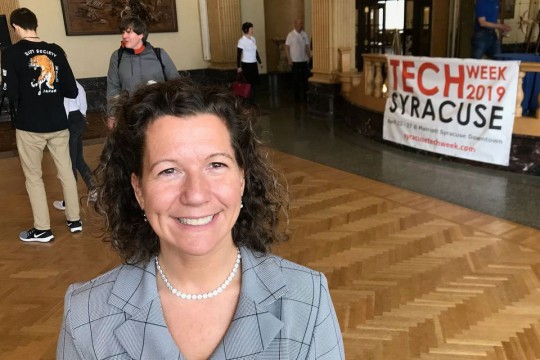 Woman poses in front of table with banner that reads: Tech Week Syracuse.