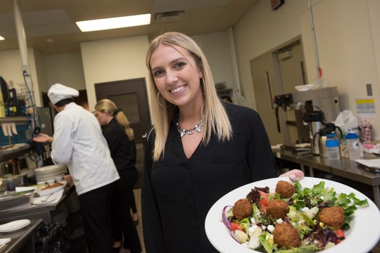 Student stands in restaurant kitchen holding plate of salad.
