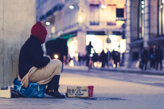 Homeless person sits at street corner.