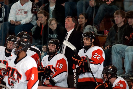 hockey players on bench with coach behind them and crowd behind him.