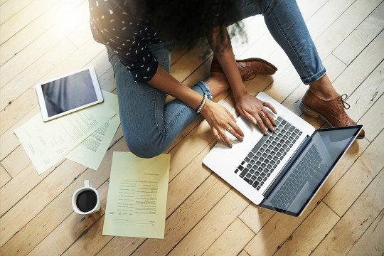 Woman sits on wood floor typing on laptop with coffee. 