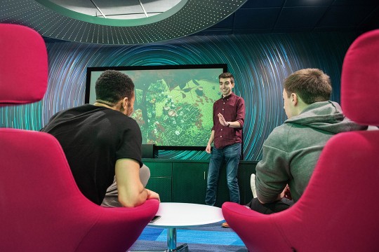 student giving presentation to two seated students while standing in front of large TV screen.