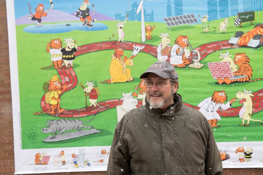 Leigh Rubin standing outdoors in front of large poster of comic.