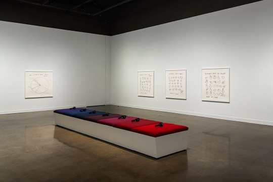 art exhibit of 7 headphones sitting on a bench with purple, blue, and red cushions.