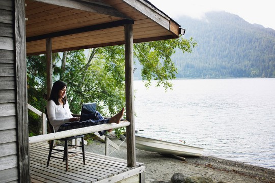 woman sitting on a deck near a lake working on a laptop.