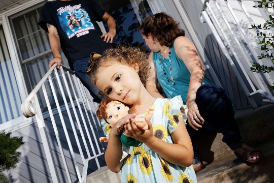 father, mother, and child holding a doll standing and sitting on the front steps to a house.