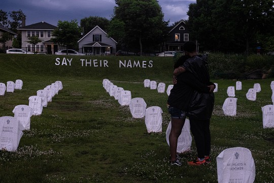two people hugging in field with makeshift grave markers and a sign that reads "Say their names."