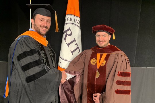 student and professor dressed in graduation regalia shaking hands.