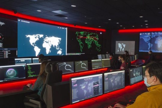 people working at table with rows of computers and large screens along the walls displays maps of the world.