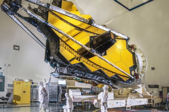 Four scientists in clean suits examine the folded James Webb Space Telescope