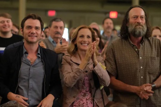 scene from a movie with three characters sitting with an audience, clapping.