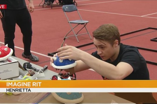 A male student makes adjustments to a robot with a blue spiky shell on top and text is overlaid that says, "IMAGINE RIT HENRIETTA"