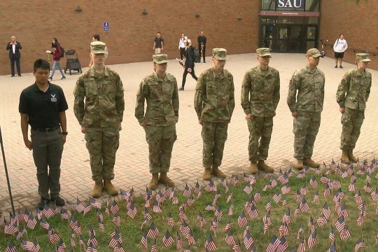seven ROTC students standing in front of a grassy area planted with small American flags.