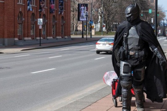 a person dressed as Batman walking down the sidewalk of a city street pulling a wagon.