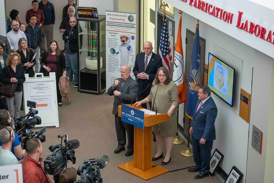 four people standing near a podium addressing a crowd including T V cameras.