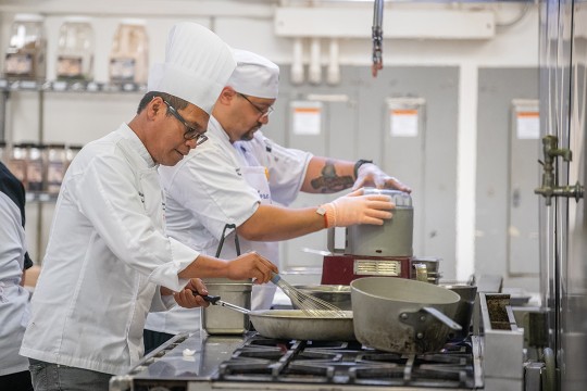 Two chef's work next to each other in a kitchen at the stove top. The first chef uses a whisk to stir food in a skillet, while the other uses a food processer.