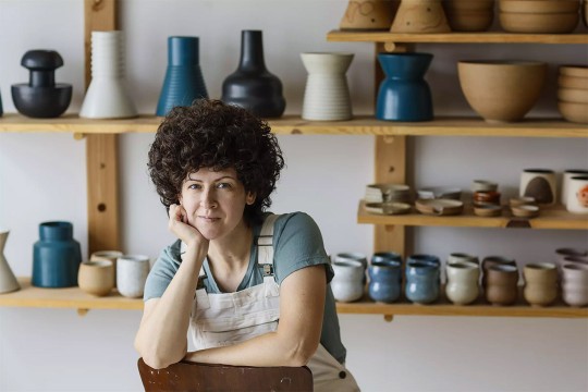 a ceramicist seated on a backwards chair, with pieces of pottery cups, bowls, and vases behind her on shelves. 