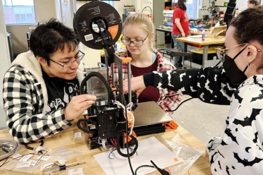 three college students working with a tabletop 3 D printer.