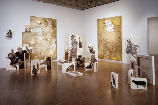In a warmly lit gallery, canvases depicting figures and animals are arrayed on the floor, with a few large-scale paintings mounted on the walls.