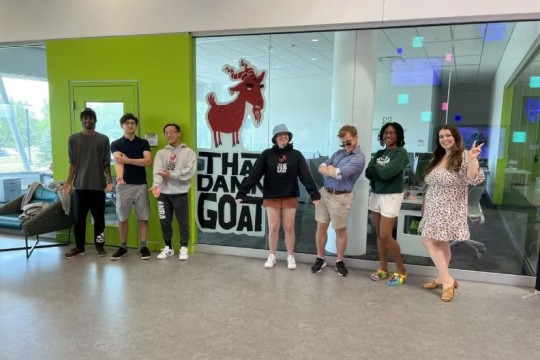 seven college student posing in front of a glass wall with a picture of a cartoon goat and the words That Damn Goat.