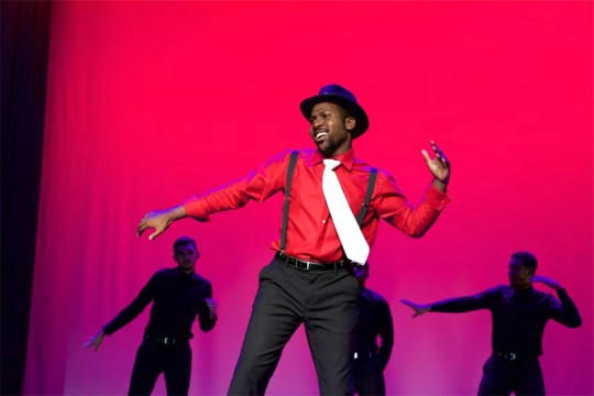 Keith Banks is shown in a red shirt with black suspenders and white tie dancing in front of a pink background