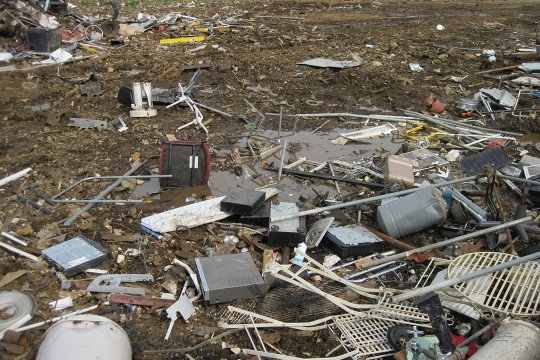 a picture of a metal scrap yard with several VCRs and DVD players sitting in the mud in the foreground that should be handled as e-waste.