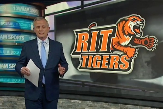 the RIT Mens Hockey athletics logo is displayed behind a newsman