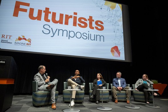 '5 people are shown on a stage, sitting in gray chairs. The speaker on the far left is holding a microphone speaking to the others. A poster that says Futurists Symposium is shown behind them.'