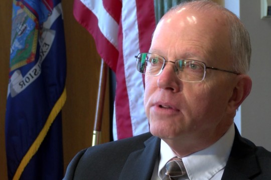 a closeup of David Munson sitting in front of the NYS and US flags.