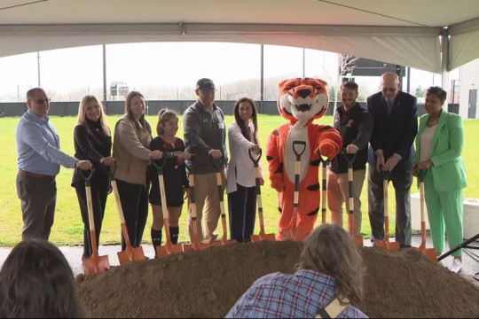 9 people and RITs mascot Ritchie stand under the shade of a tent in a line with shovels in front of a mound of dirt marking the groundbreaking of the new stadium.