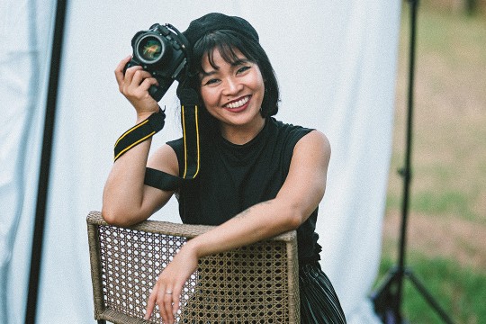 Joi Conti sits in a wicker chair that is facing backwards. She is wearing a sleeveless black top, a black hat, and is holding a camera near her head.