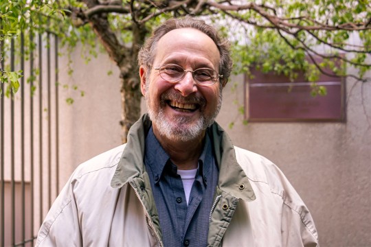 Leigh Rubin is shown in a tan jacket standing in a courtyard smiling with glasses on.