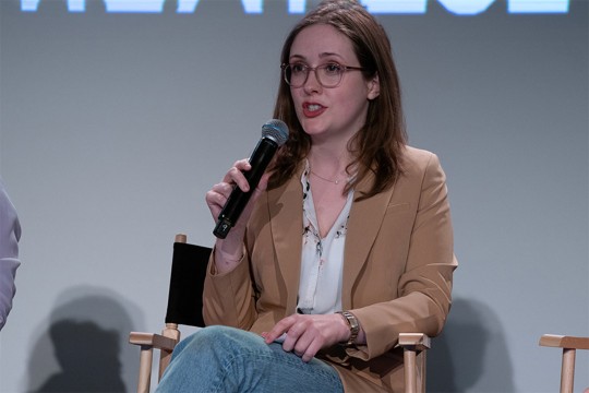 Molly HIll is shown sitting on a directors style chair in jeans and a light brown suit jacket.