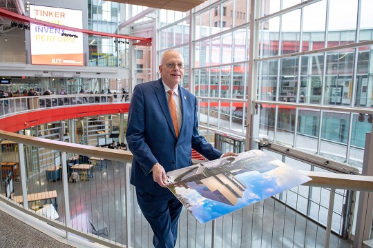 'David Munson, RIT President, stands on the second floor balcony of the SHED holding a portrait that shows the renderings before it was built.'