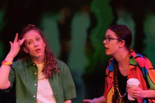 two women appear on stage for a play at NTID. The woman on the left is in a green shirt holding her hand to her head and the woman on the right appears in to be speaking to the other woman while wearing a brightly colored shirt and holding a coffee.