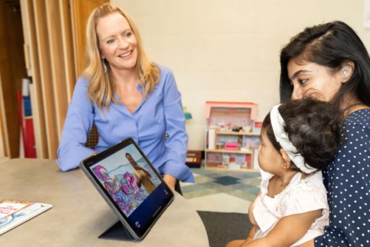 'Rain Bosworth smiling and looking at a parent-child pair to her left. She has blonde hair and blue eyes and wearing blue button-up shirt. The parent is looking at an iPad, sitting in front of them on a round table. The iPad is displaying what appears to be a video with a person signing. The parent has black hair and wearing a navy polka dot shirt. The child is sitting on the parent's lap and staring at Bosworth.'