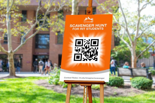 an orange and white sign reading Scavenger Hunt with a QR code is shown on an easel sitting in a common area outside in the academic space on campus.