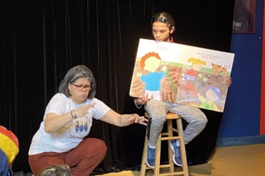 a woman is shown signing for a student on a stage who is sitting on a stool and holding an open book for display. 