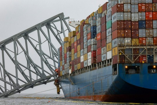 'a large ship carrying cargo containers is shown with a partial piece of the baltimore bridge hanging off the side.'