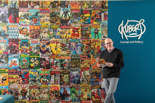 a man stands next to a wall covered in comic book covers. On the right side of the wall there is a sign that says Kubert Lounge and Gallery.