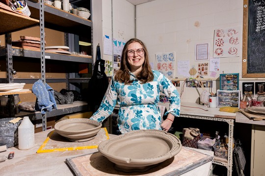 a college age girl stands in a ceramic studio room with two large, raw clay serving platters in front of her.