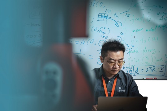 a man looks at a computer laptop screen while sitting in front of a whiteboard with formulas scribbled across it.