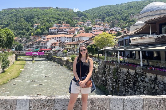 'a college age girl is shown standing on a bridge overlooking a town in Kosovo.'