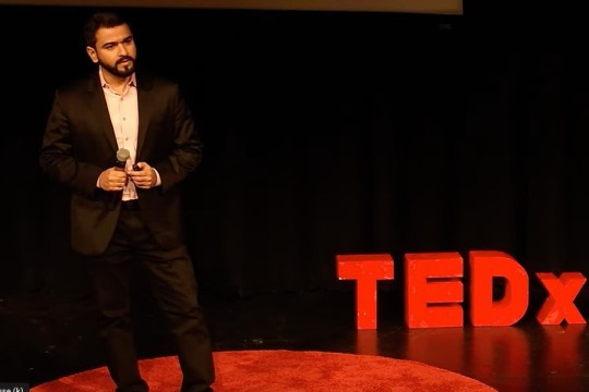 Ahmad Kirmani stands on a red circle rug in front of a black background. A sign reading TEDx appears next to him at floor level.