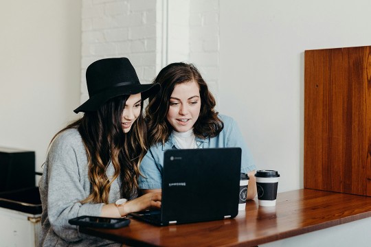 two students sit at a small table looking at a laptop together.