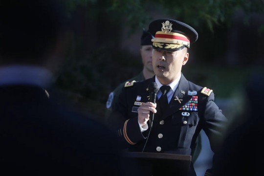a man in military dress speaks in to a microphone in an outdoor space.