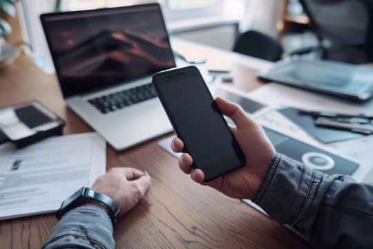 A pair of hands holds a smartphone of an unknown brand. In the background, there is a laptop and a spread of papers across the desk as though someone was in the middle of working on a project. The items in the background are slightly blurred and the main focus is on the phone, which has a black screen.