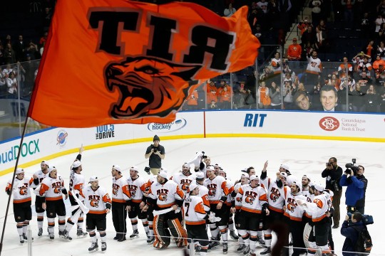 A top-down view of the RIT Hockey team celebrating a win on the ice. There is a large orange RIT flag waving in the foreground of the image, and there are a number of camera people and videographers on the ice with the team.