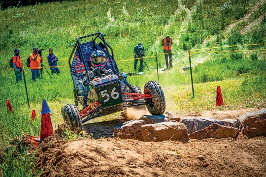 A baja car drives over a dirt road littered with obstacles.