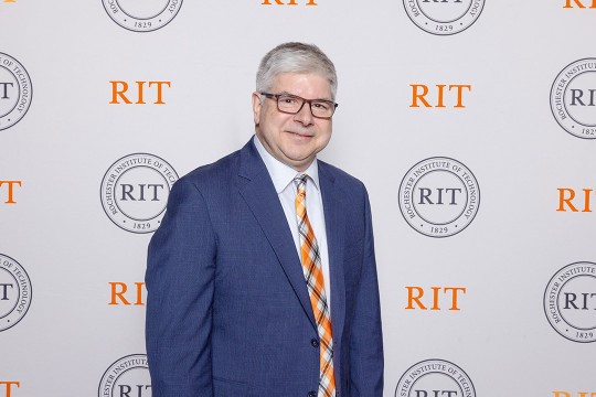 William Sanders appears in a blue suit standing in front of a step and repeat bearing RITs logos and marks.